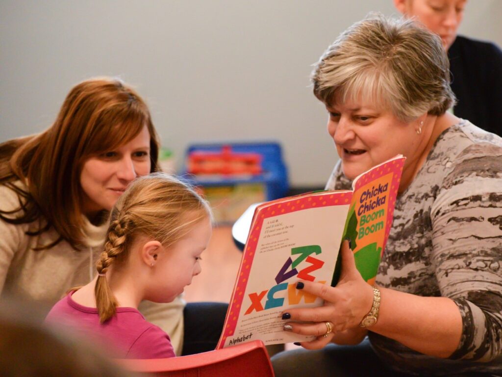A teacher reads Chicka Chicka Boom Boom to a young girl while another adult listens and smiles.