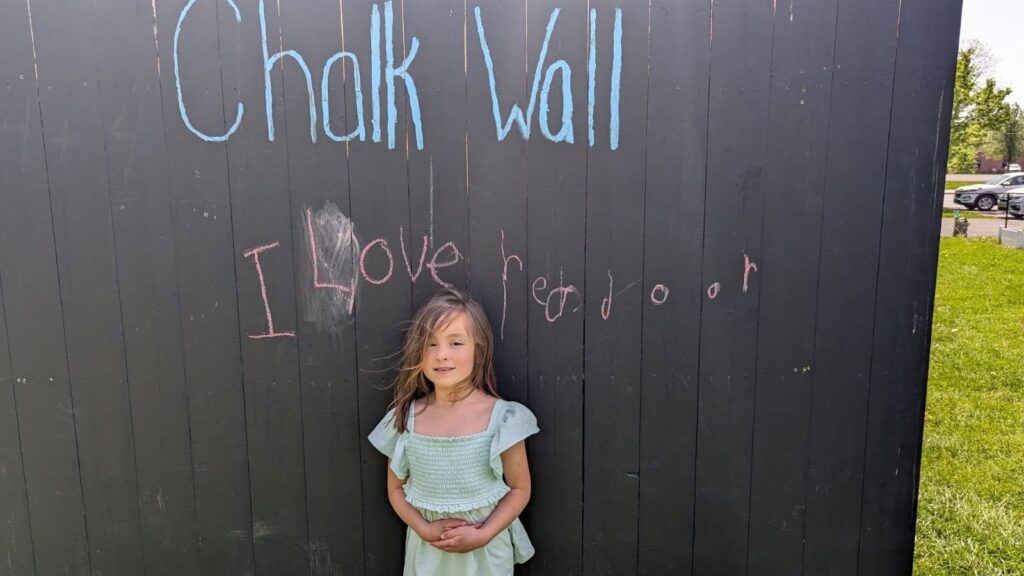A young girl stands in front of a chalkboard wall with the message “I love red door” written in chalk.