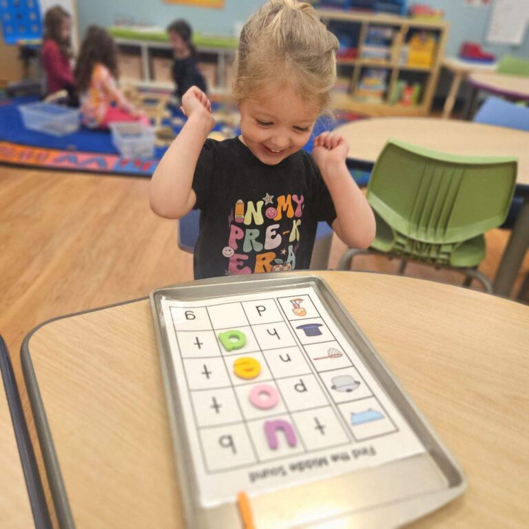 A preschooler smiles with excitement at a table activity featuring letter matching.